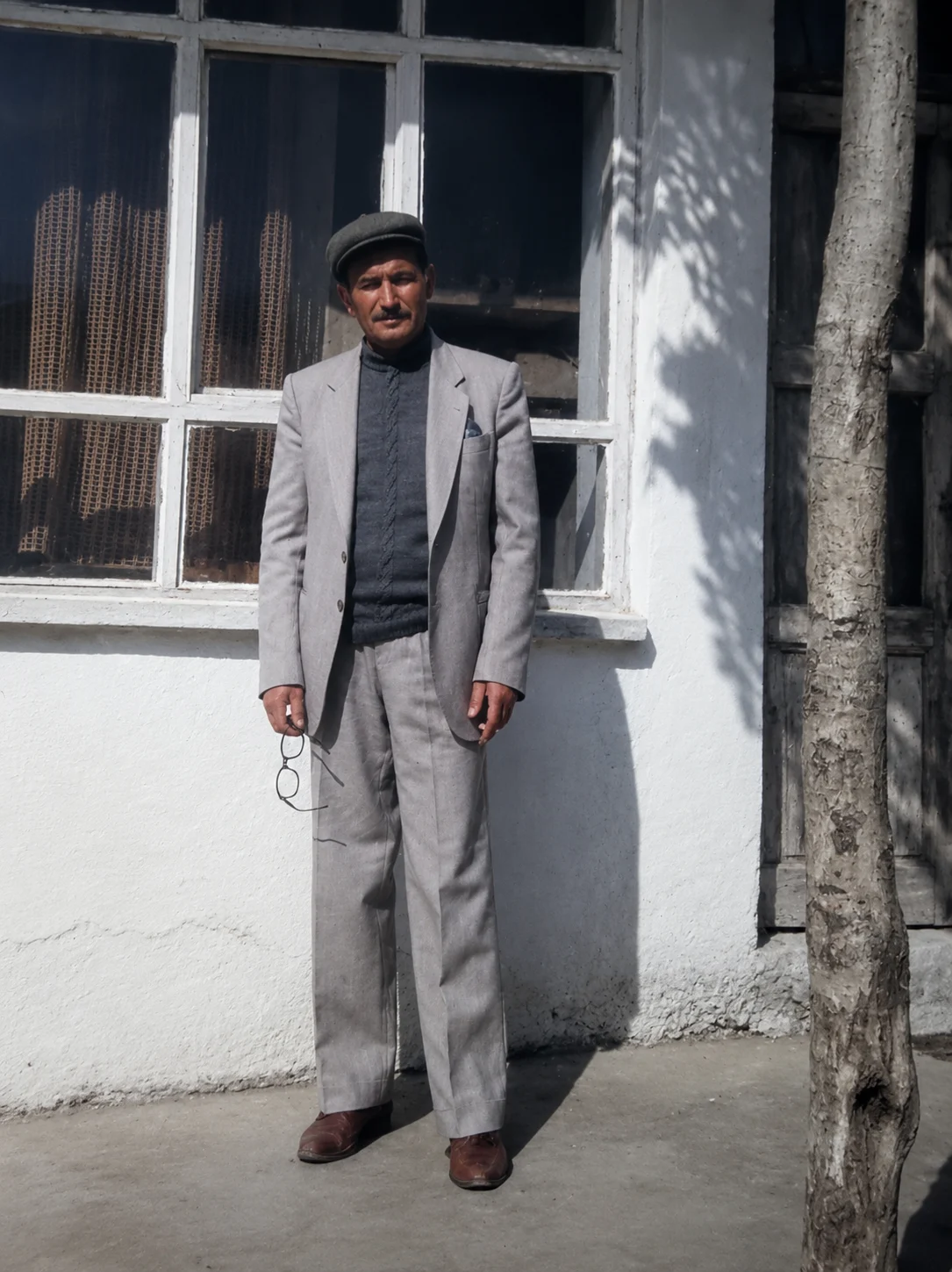 Muttalip Erdem, evinin önünde, elinde gözlüğüyle ayakta duruyor.